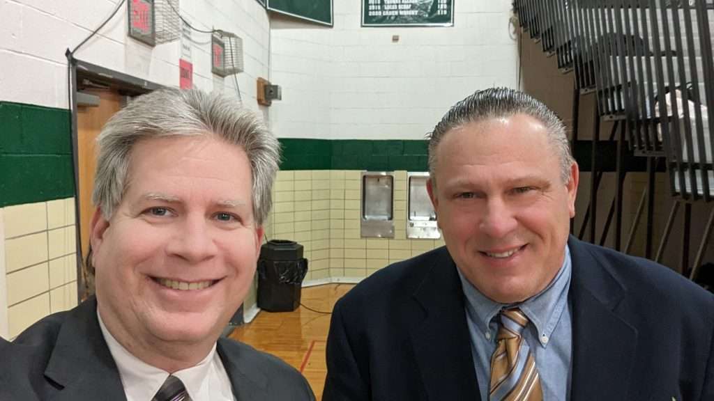 two middle aged men smiling at camera at a basketball game