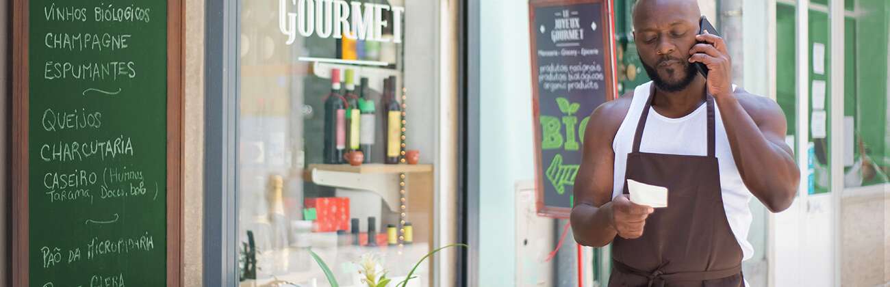 Dark-skinned man in a trendy grocer apron uses mobile phone outside a boutique gourmet shop