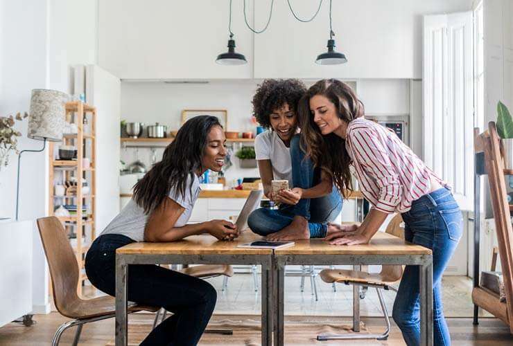 Three women with multiple mobile devices gather around a table looking at a mobile phone