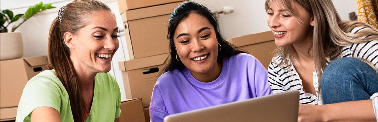 Three women set up WiFi at a new apartment using a laptop sitting on moving boxes
