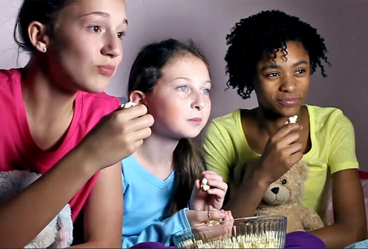 Three young multiracial girls with popcorn stare transfixed at the TV