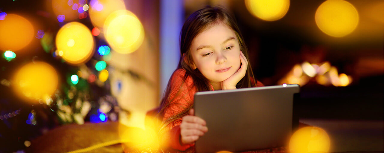 Young girl surrounded by holiday lights watches movies streaming on her tablet.