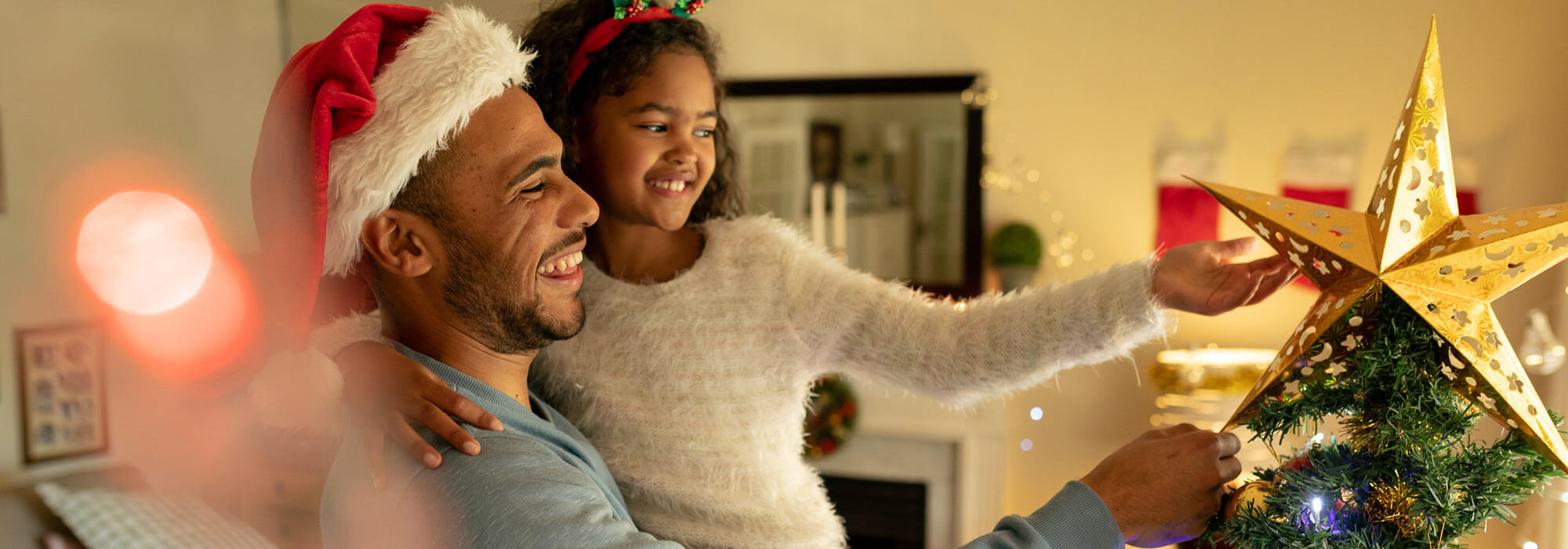 Daughter in holiday attire adds the star at the top of the Christmas tree with Dad holding her up to reach.