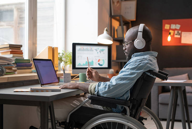 Data professional seated in a wheelchair works from home with a multi-device setup and headphones