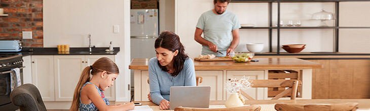 Mom on laptop helps daughter with homework at kitchen table while Dad fixes dinner