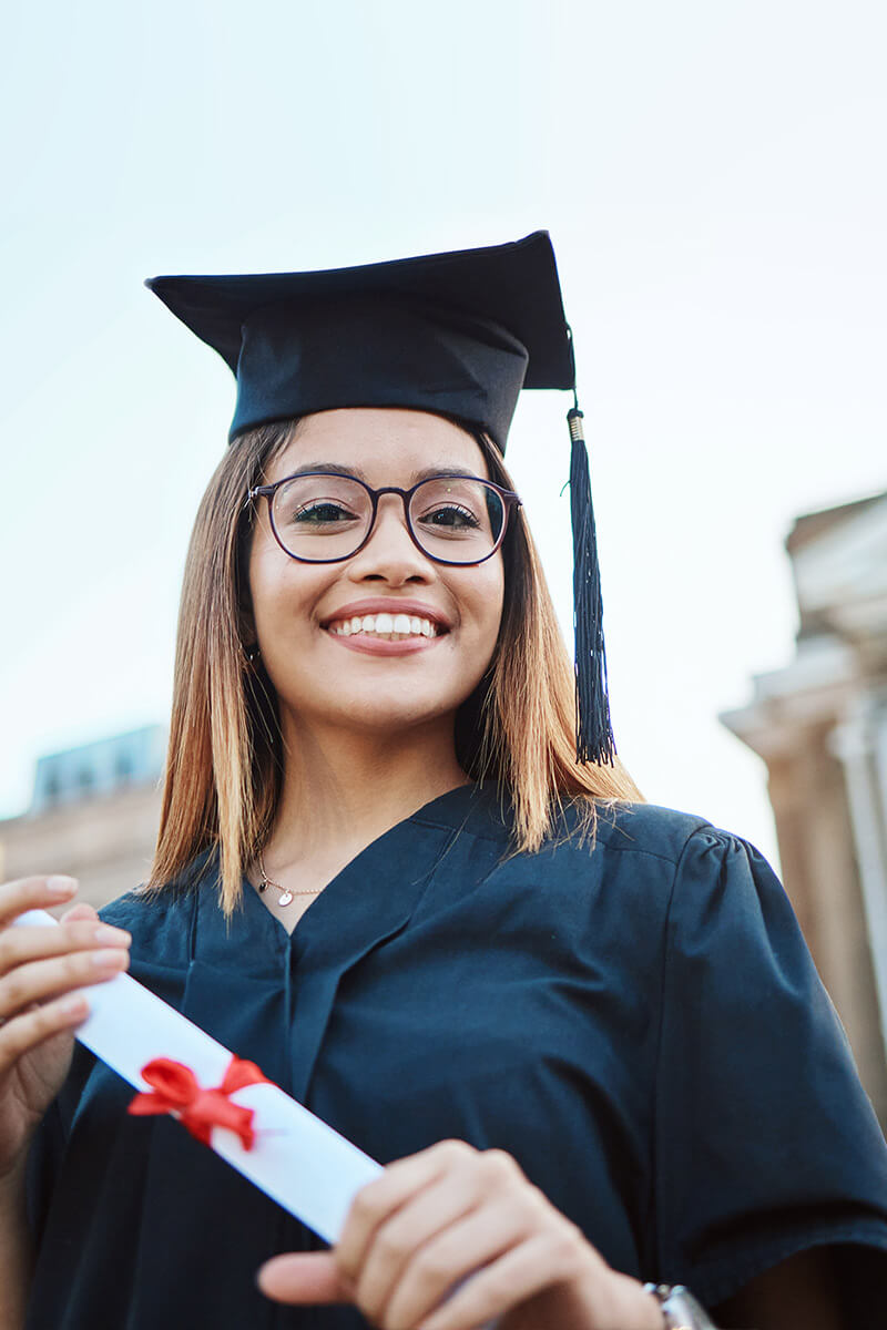young woman holds diploma in front of classical style school buildings